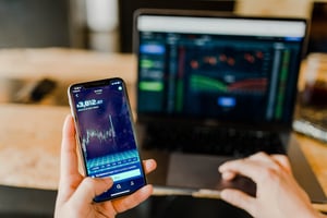 a person looking at stocks on a phone and on a laptop.